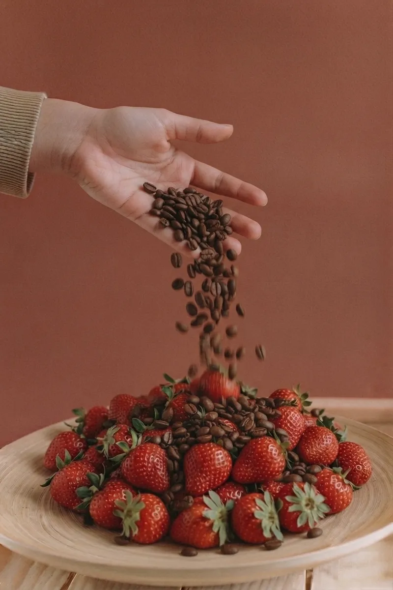 Hand pouring coffee beans over a pile of fresh strawberries